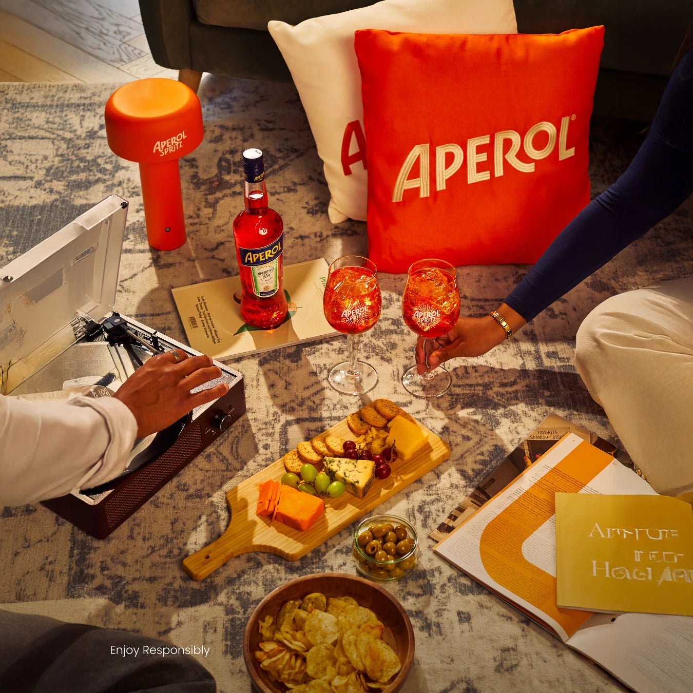 Aperol Spritz bottle, glasses and snacks on a cozy rug with pillows, record player and magazines